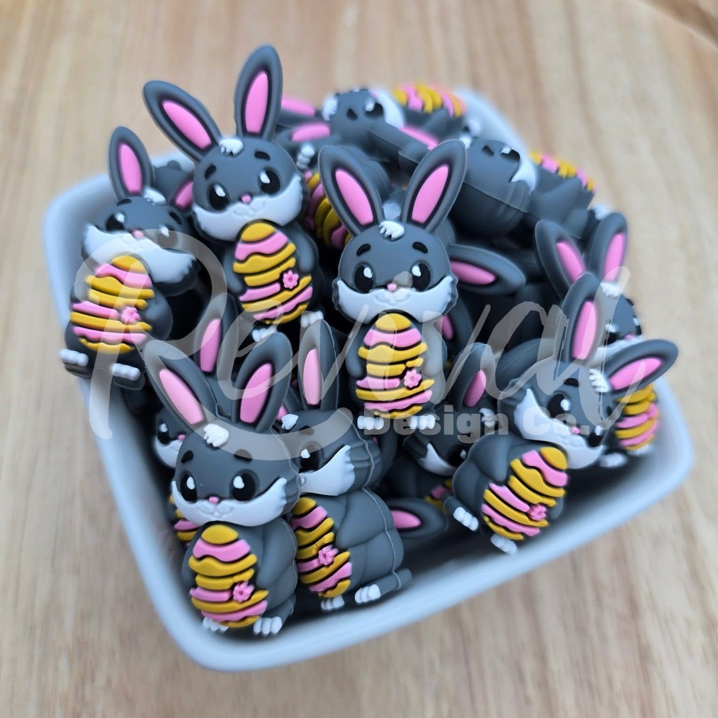 Collection of cartoonish rabbit beads holding striped easter eggs on a wooden surface, in a white ceramic bowl.