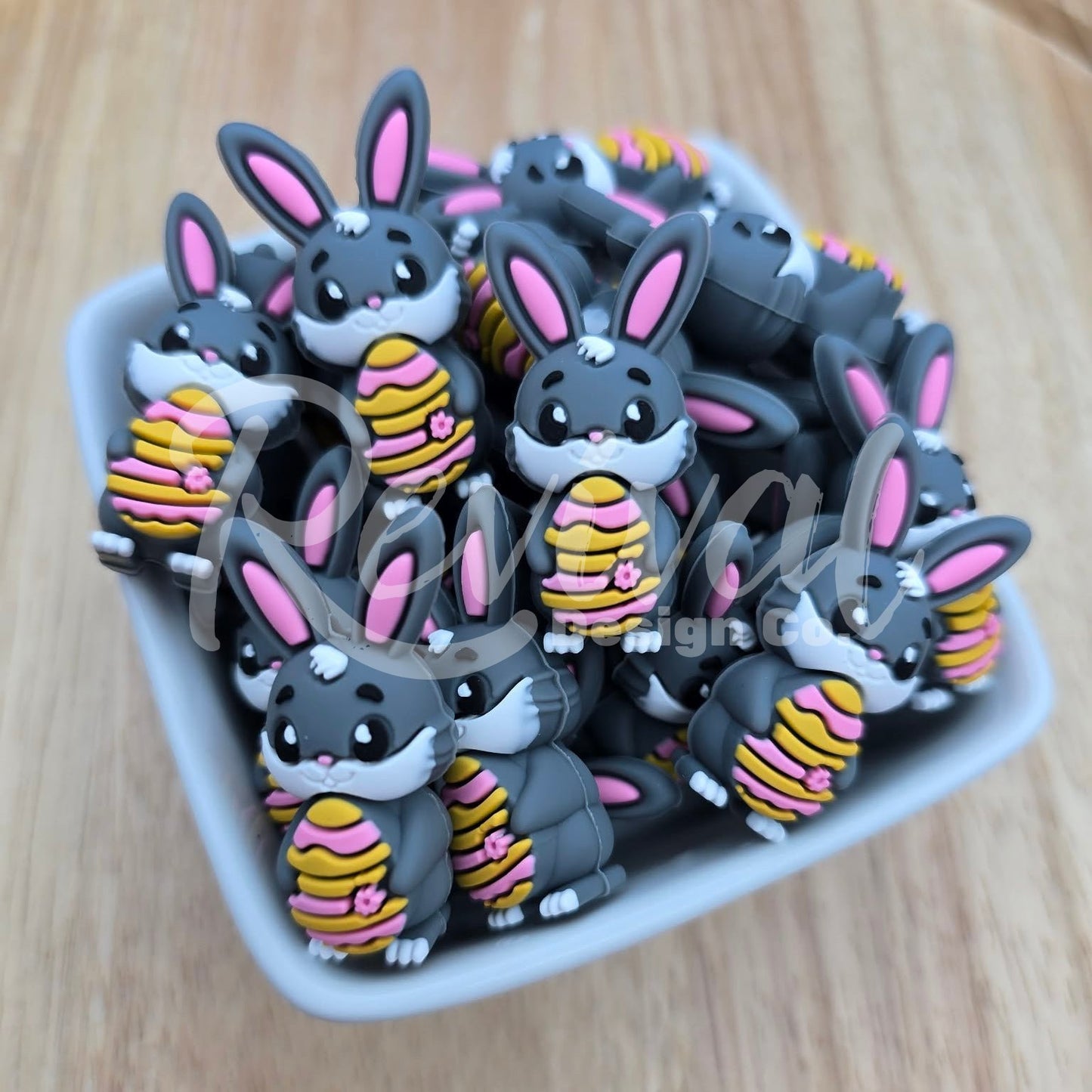 Collection of cartoonish rabbit beads holding striped easter eggs on a wooden surface, in a white ceramic bowl.