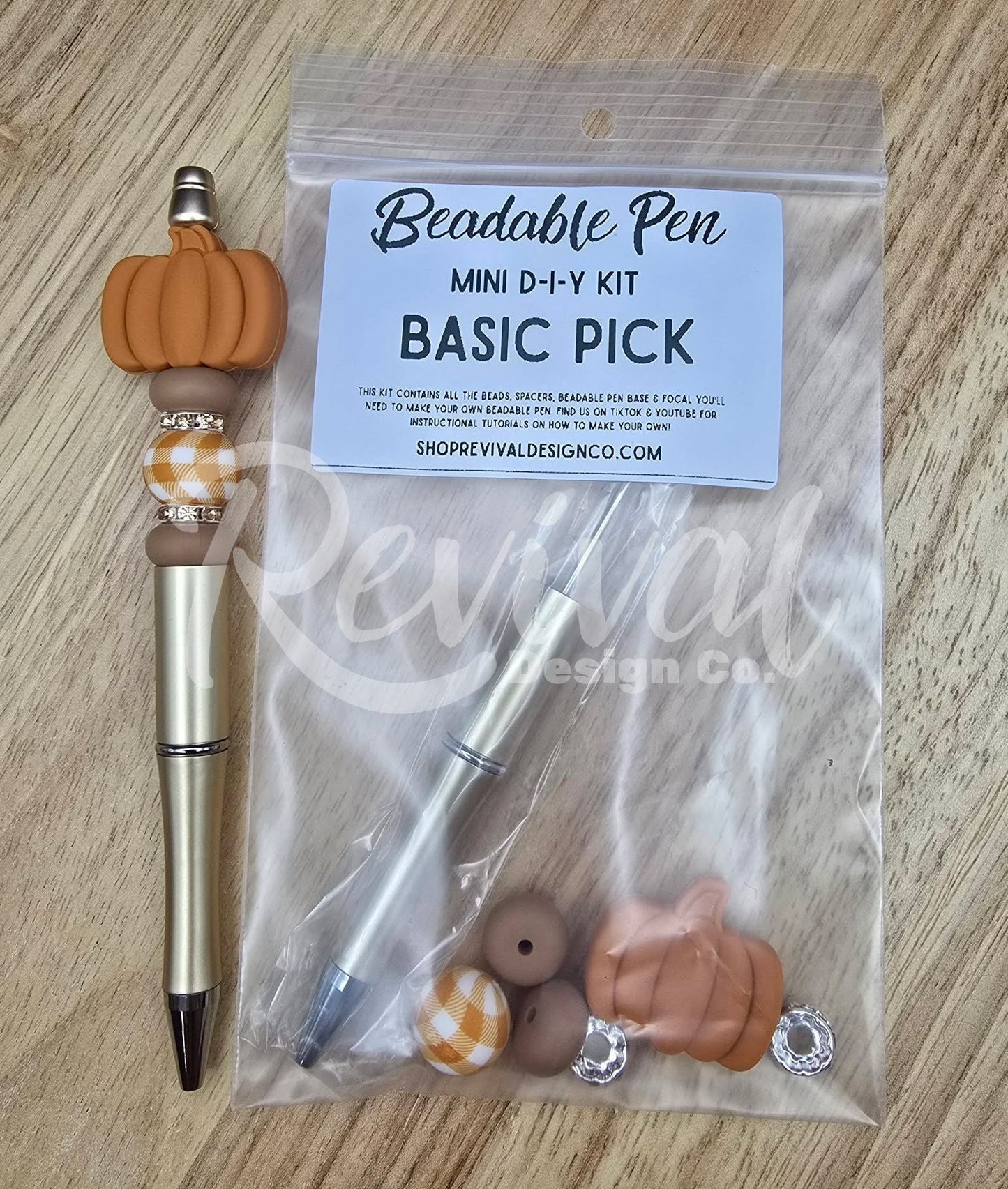 A mini DIY beaded pen kit labeled 'Beadable Pen MINI D-I-Y KIT BASIC PICK' in a clear plastic bag, shown next to a completed example pen with a metallic gold body, black tip, and orange pumpkin charm on a light wood surface.