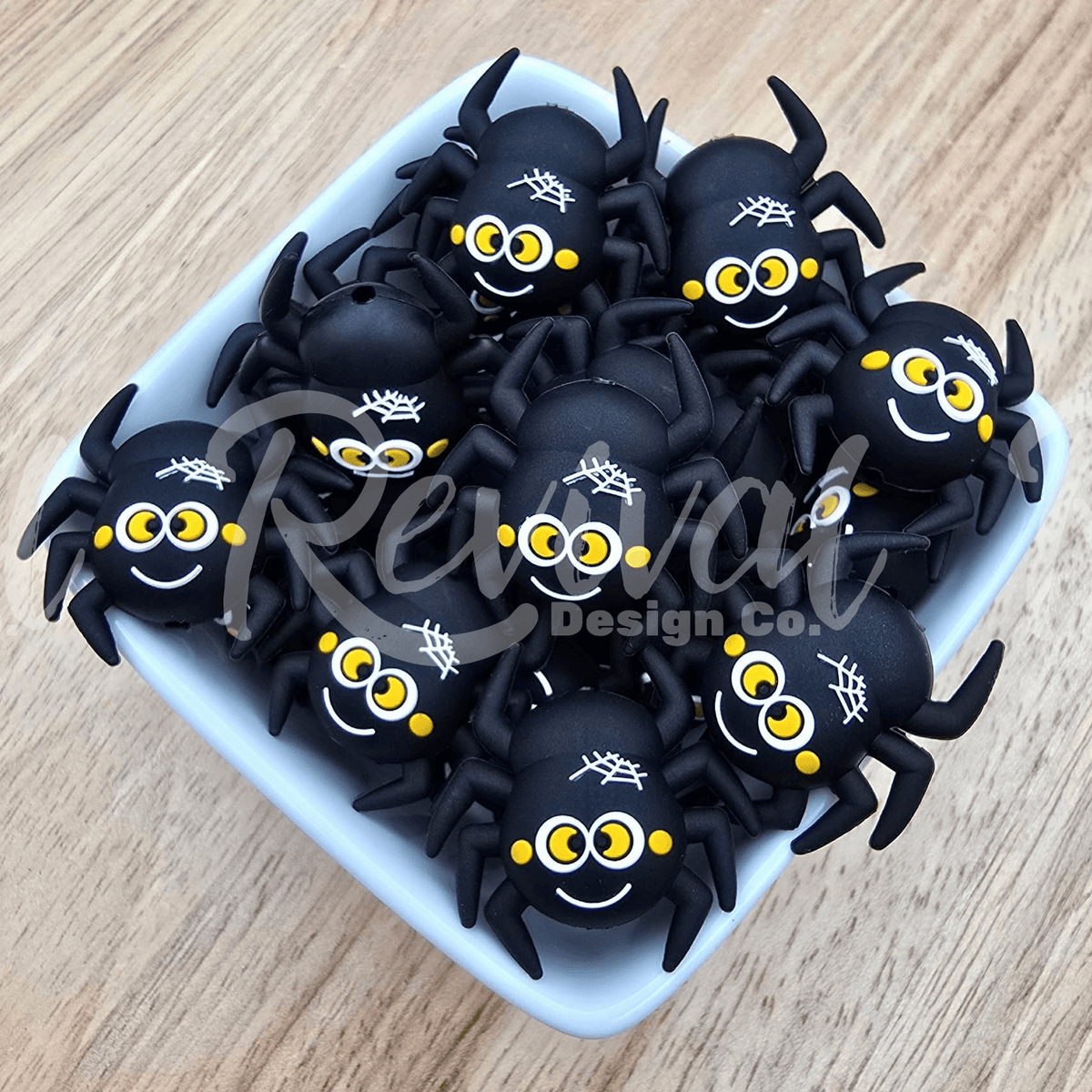A group of black plastic spider-shaped items with yellow eyes, white spiderwebs, and smiling faces, arranged in a small white square dish on a light wood surface.