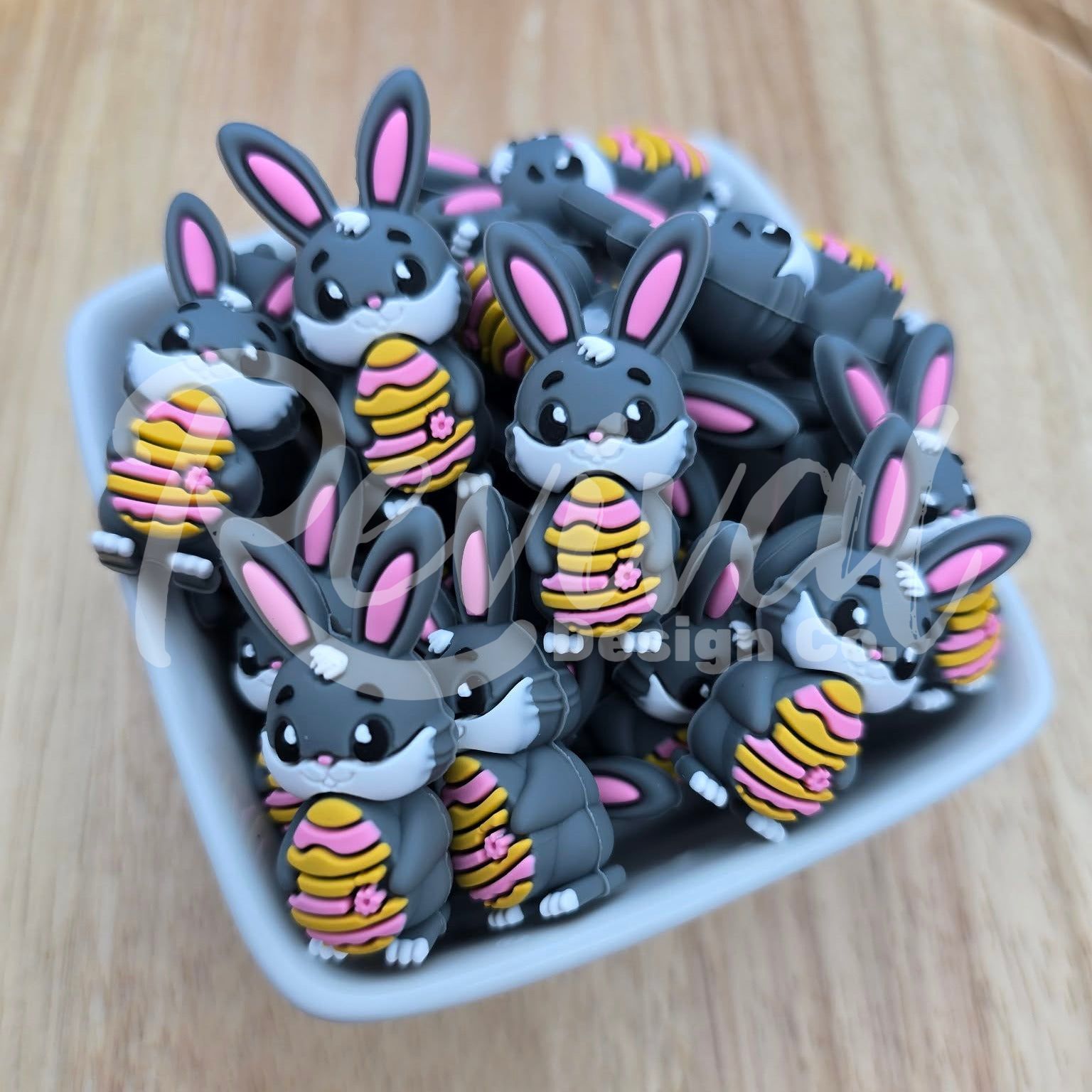 Collection of cartoonish rabbit beads holding striped easter eggs on a wooden surface, in a white ceramic bowl.