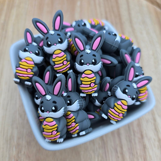 Collection of cartoonish rabbit beads holding striped easter eggs on a wooden surface, in a white ceramic bowl.