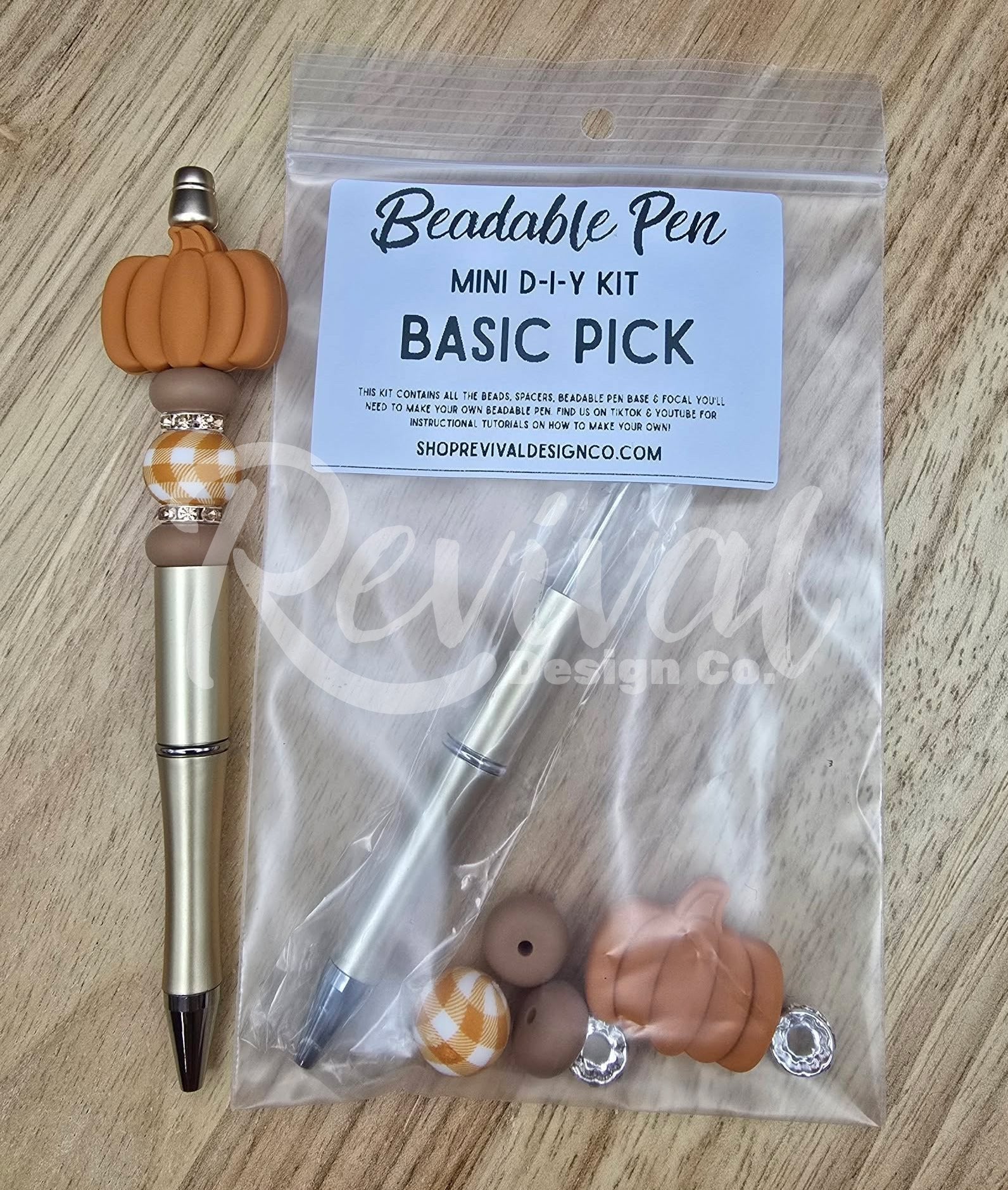 A mini DIY beaded pen kit labeled 'Beadable Pen MINI D-I-Y KIT BASIC PICK' in a clear plastic bag, shown next to a completed example pen with a metallic gold body, black tip, and orange pumpkin charm on a light wood surface.