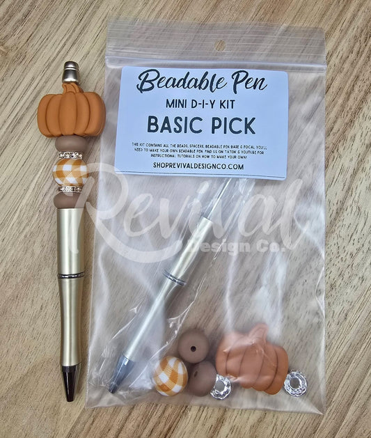 A mini DIY beaded pen kit labeled 'Beadable Pen MINI D-I-Y KIT BASIC PICK' in a clear plastic bag, shown next to a completed example pen with a metallic gold body, black tip, and orange pumpkin charm on a light wood surface.