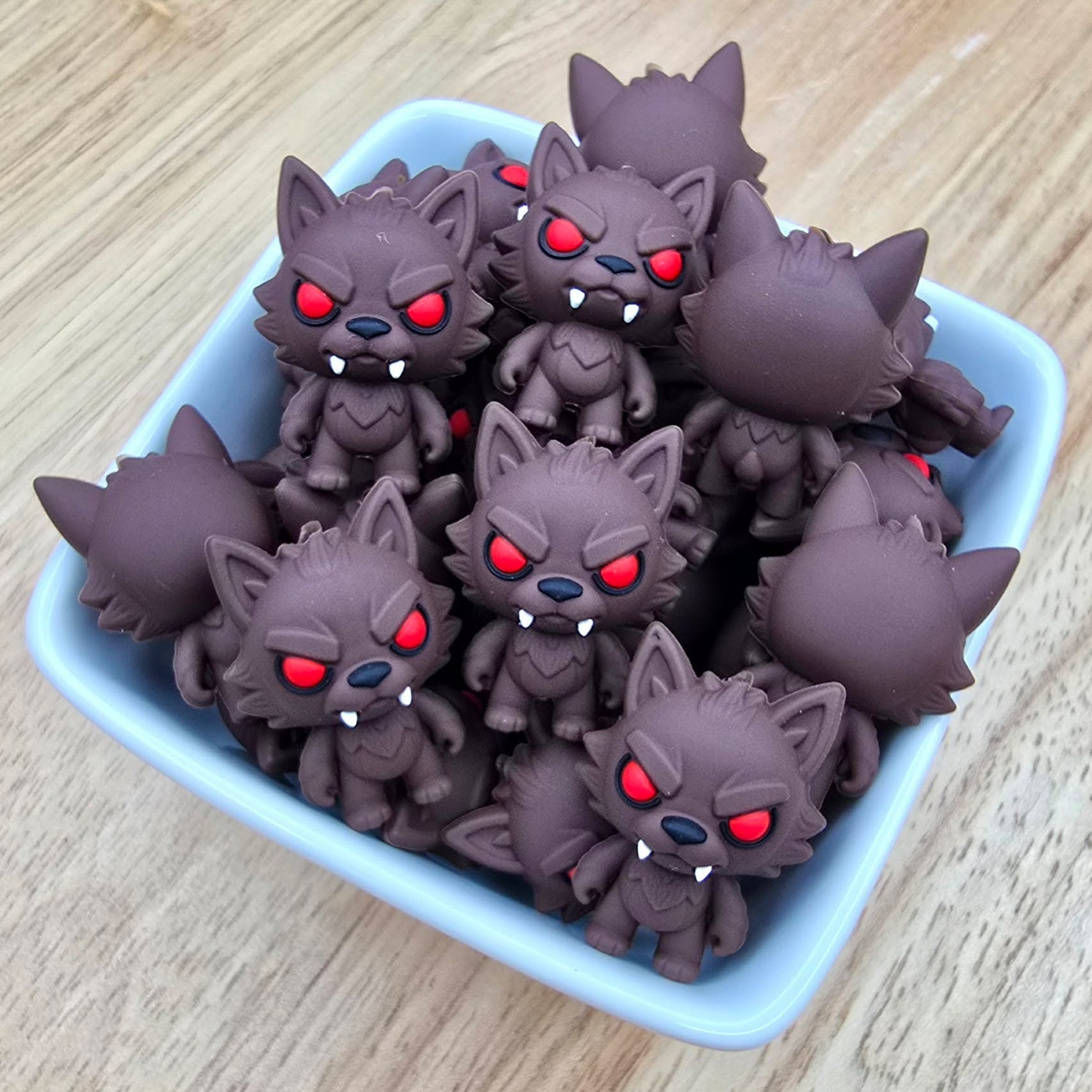 A group of small dark brown cartoon wolf figurines with glowing red eyes and white fangs, arranged in a light blue square dish on a light wood surface.