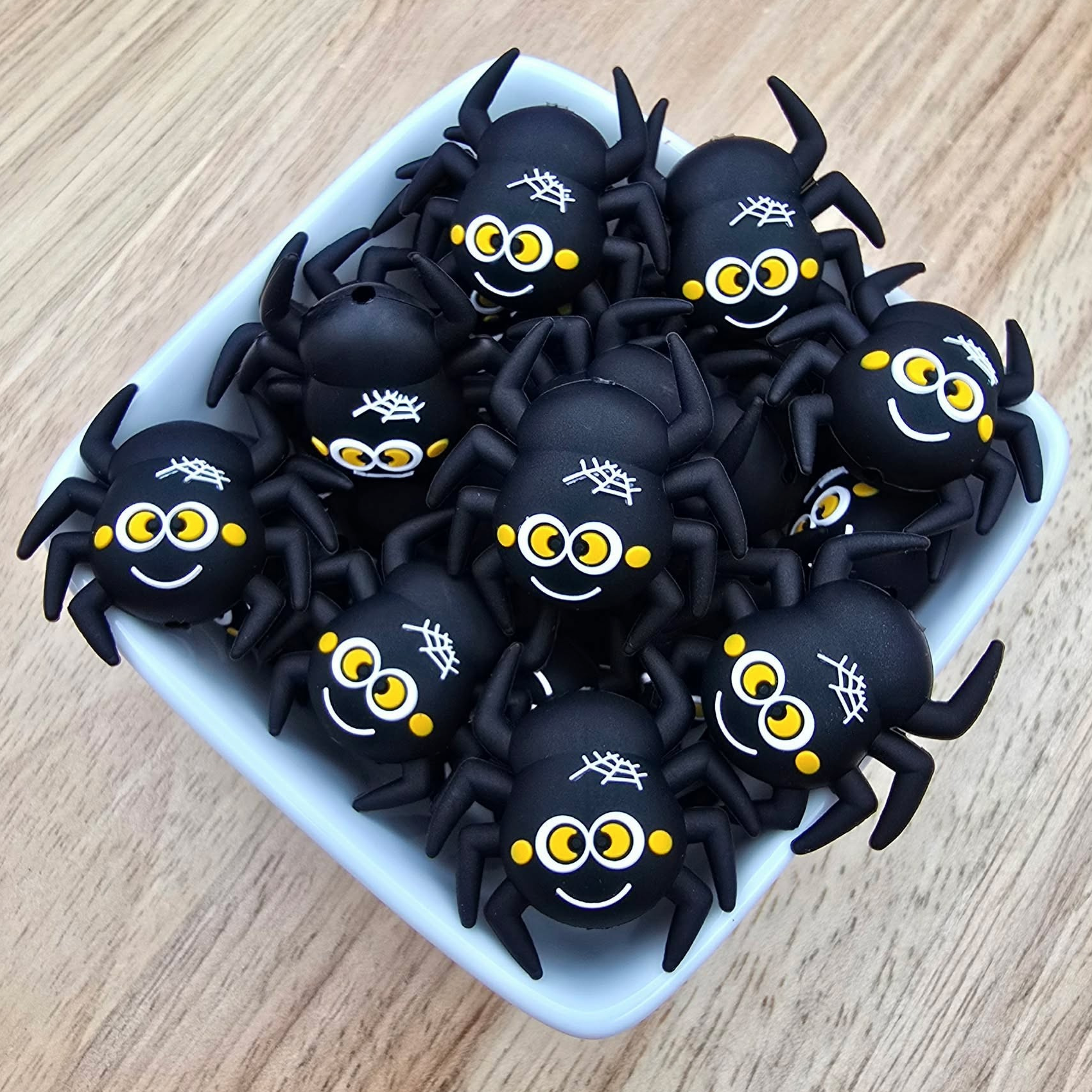 A group of black plastic spider-shaped items with yellow eyes, white spiderwebs, and smiling faces, arranged in a small white square dish on a light wood surface.